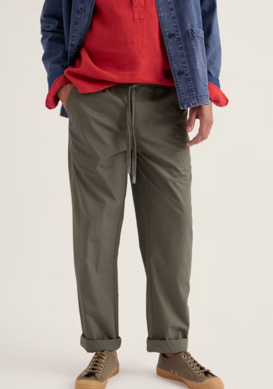 Seasalt Men's Kenython Relaxed Tapered Trousers Light Nori green that are relaxed tapered leg with Elasticated waist with drawstring tie and a Concealed zip fastening with Side pockets and Back pocket that's Machine Wash 30C Material: 100% Organic Cotton Person wearing a red shirt, blue jacket, and green pants on a white background