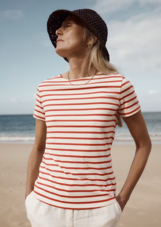 Seasalt Sailor T-Shirt Breton Carnelian. Womens tee that's Semi fitted and Yarn-dyed stripes with Boat neck that's Machine Wash 30C Material: 100% Organic Cotton Woman wearing a red and white striped shirt and black hat on a beach.