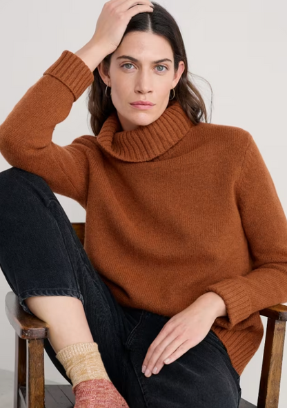 Seasalt Braque Lambswool Blend Roll Neck Jumper Rusted Seasalt clothing womens wool jumper Woman wearing a brown sweater sitting on a wooden chair against a white background