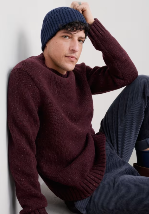 Seasalt Men's Bizen Jumper Merlot Mens Jumper Mens Wool Jumper Seasalt clothing Man wearing a maroon sweater and blue beanie sitting against a light gray wall.