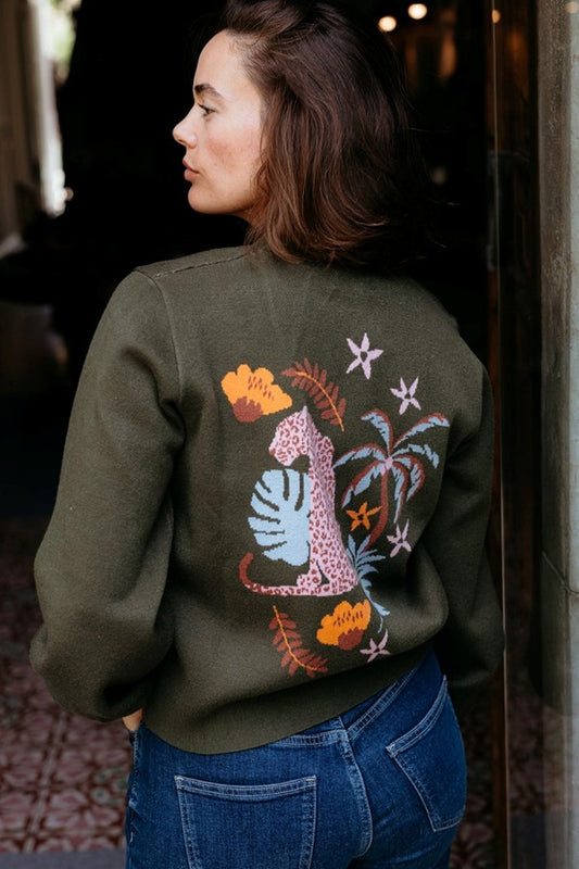 Sugarhill Brighton Adrian Cardigan Khaki Leopard Jungle womens Buttoned front with V-neck and Blouson sleeve that's Machine washable (delicate) Colour: Khaki Leopard Jungle Material: 51% Viscose, 30% Polyester & 19% Nylon Person wearing a green sweatshirt with colorful designs on the back, standing indoors.