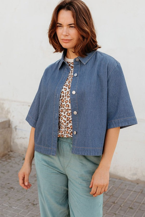 Sugarhill Brighton Amity Jacket Blue Stripe Denim womens Button down front that's Mid-length, wide sleeves and a Shorter boxy shape that's Classic shirt collar and Machine washable (delicate) Colour: Blue Stripe Denim Material: 100% Responsibly Sourced Cotton Woman wearing a blue jacket over a leopard print top and green pants against a white wall.