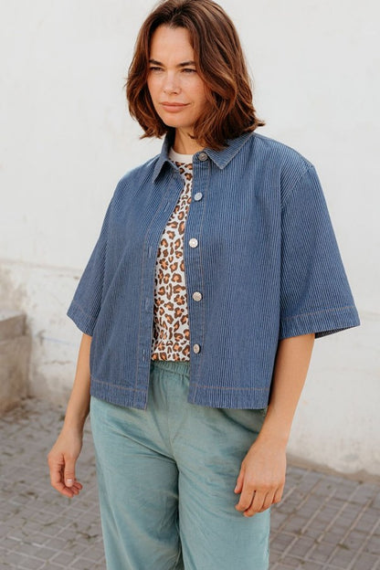 Sugarhill Brighton Amity Jacket Blue Stripe Denim womens Button down front that's Mid-length, wide sleeves and a Shorter boxy shape that's Classic shirt collar and Machine washable (delicate) Colour: Blue Stripe Denim Material: 100% Responsibly Sourced Cotton Woman wearing a blue jacket over a leopard print top and green pants against a white wall.