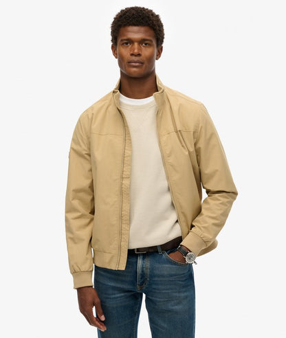 Superdry Nylon Harrington Jacket Canyon Beige mens in a Relaxed fit has a Zip fastening and Ribbed cuffs and hem with Two waist pockets and internal pocket that's Fully lined Rubberised logo on sleeve polyamide & Recycled Polyester Canyon Beige Style Code: M5012079A Man wearing a beige jacket over a white shirt with blue jeans on a white background