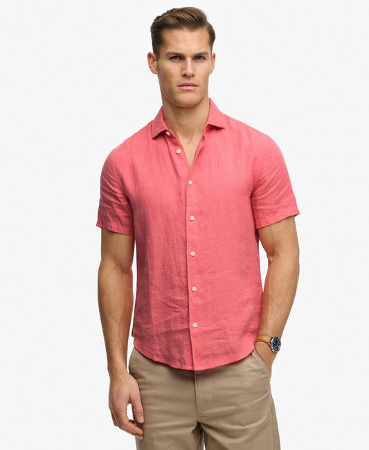 Superdry Riviera Short Sleeve Linen Shirt Coral mens that has a Relaxed fit Embroidered logo on the chest a Standard collar that's Button fastening with Short sleeve and Signature tab on the hemline Colour: Coral Material: 100% Linen Man wearing a pink short-sleeve shirt and beige pants on a white background