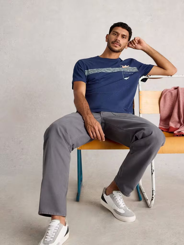 White Stuff Abersoch Tee Graphic In The Ocean Navy Print mens T-shirt with Crew Neck and Short Sleeve that's Regular Fit and Machine Washable Colour: Navy Print Material: 100% Cotton Man sitting on a chair wearing a blue t-shirt and gray pants, with a neutral background.
