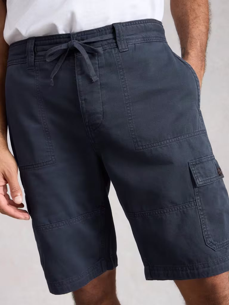 White Stuff Kemble Cargo Shorts Dark Navy. Men's Classic Fit Shorts that are Machine Washable in Dark Navy Material: 100% Organic Cotton. Crafted from organic cotton, these White Stuff Kemble cargo shorts deliver comfort and durability for summer days Navy cargo shorts worn by a person on a plain background