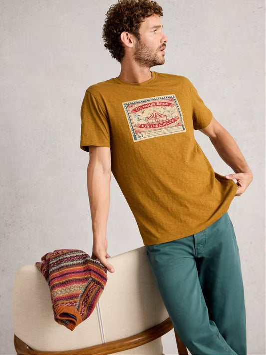 White Stuff Abersoch Graphic Circus Yellow Man wearing a mustard yellow t-shirt with a graphic design, standing next to a chair with a colorful blanket.
