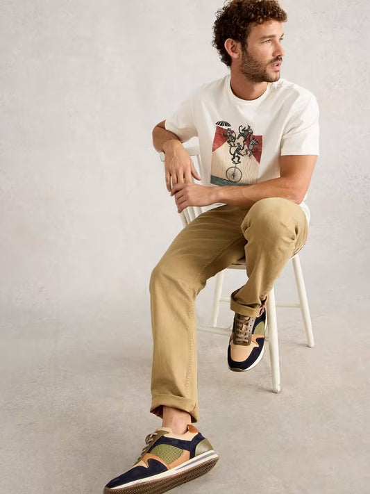 White Stuff Abersoch Graphic Monkey White Man sitting on a chair wearing a white t-shirt, beige pants, and patterned shoes.