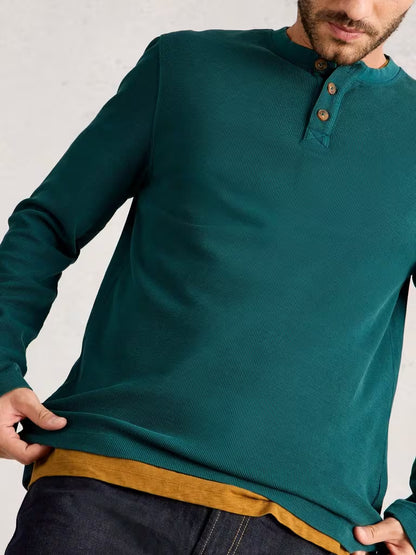 White Stuff Helston Waffle Henley Mid Green Man wearing a teal long-sleeve shirt with brown trim against a light gray background