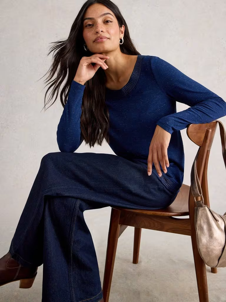 White Stuff Weaver LS Sparkle Tee Dark Navy Womens Jumper White Stuff Clothing Woman sitting on a wooden chair wearing a blue sweater and dark pants, with a beige handbag beside her.